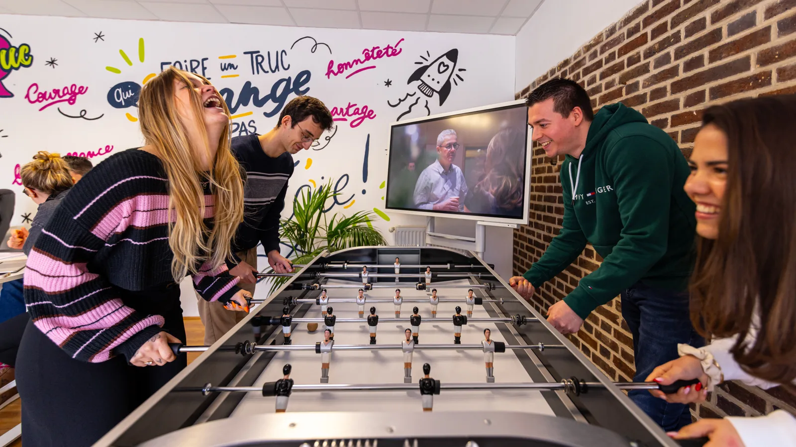L’équipe Zetruc en pleine partie de baby-foot à l’agence, devant le mur peint aux mots de la marque.